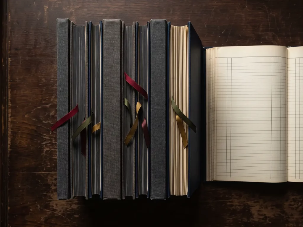 Cenital de tres pilas de libros de cuentas con cintas marcapáginas sobre madera oscura, junto a un libro abierto de rayado en blanco.